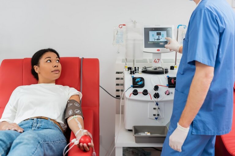 doctor in blue uniform and latex gloves operating transfusion machine near multiracial woman sitting